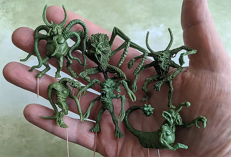 A hand holds several green resin miniatures, showcasing a variety of fantastical creatures. The figures include grotesque designs with multiple limbs, horns, and exaggerated features, reflecting an old-school aesthetic reminiscent of classic tabletop miniatures. Each miniature is mounted on a small pin for display, emphasizing their intricate details and unique forms.