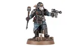 A detailed miniature figure of a Death Korps of Krieg Combat Engineer stands on a textured base, holding a hammer in one hand and a pistol in the other. The figure is dressed in dark, militaristic attire with a distinctive gas mask and helmet, featuring various armor plates and insignia. The base includes small wooden debris, enhancing the battlefield aesthetic.