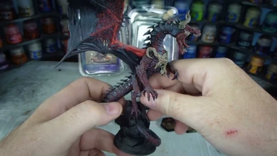 A detailed dragon miniature is being held in a hand, showcasing its intricate features, including prominent scales, fierce facial expression, and large wings with a red underside. The dragon is painted in dark hues with highlights that accentuate its muscular form and menacing appearance. In the background, various paint jars are visible, indicating a workspace for model painting.