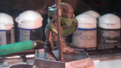 A small, detailed miniature figure stands on a square base, holding a staff and wearing a green mask. The figure is dressed in textured armor, showcasing intricate details like scales and straps. In the background, various paint pots and tools are visible, indicating a workspace for painting or modeling miniatures.