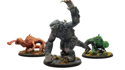 The image features three miniatures from Modiphius’ Fallout: Wasteland Warfare line. On the left is a snallygaster, a hexapod creature with a reddish-brown body, multiple eyes on its back, and a long, protruding tongue. In the center stands a large, muscular megasloth, covered in gray fur, with a menacing pose and a raised arm. To the right is another creature, possibly a variant of the snallygaster, in green, with a textured body and large eyes, all set on circular bases adorned with tufts of grass.
