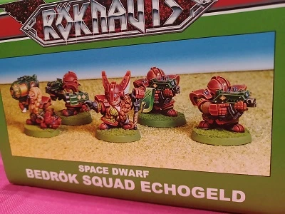 A box labeled "ROKNAUTS" features a colorful illustration of five miniatures, known as the Bedrök Squad Echogeld, designed as space dwarves. Each miniature is painted in vibrant colors, showcasing intricate details such as armor and weaponry, set against a sandy backdrop. The box design includes bold graphics and text, emphasizing the theme of the miniatures.