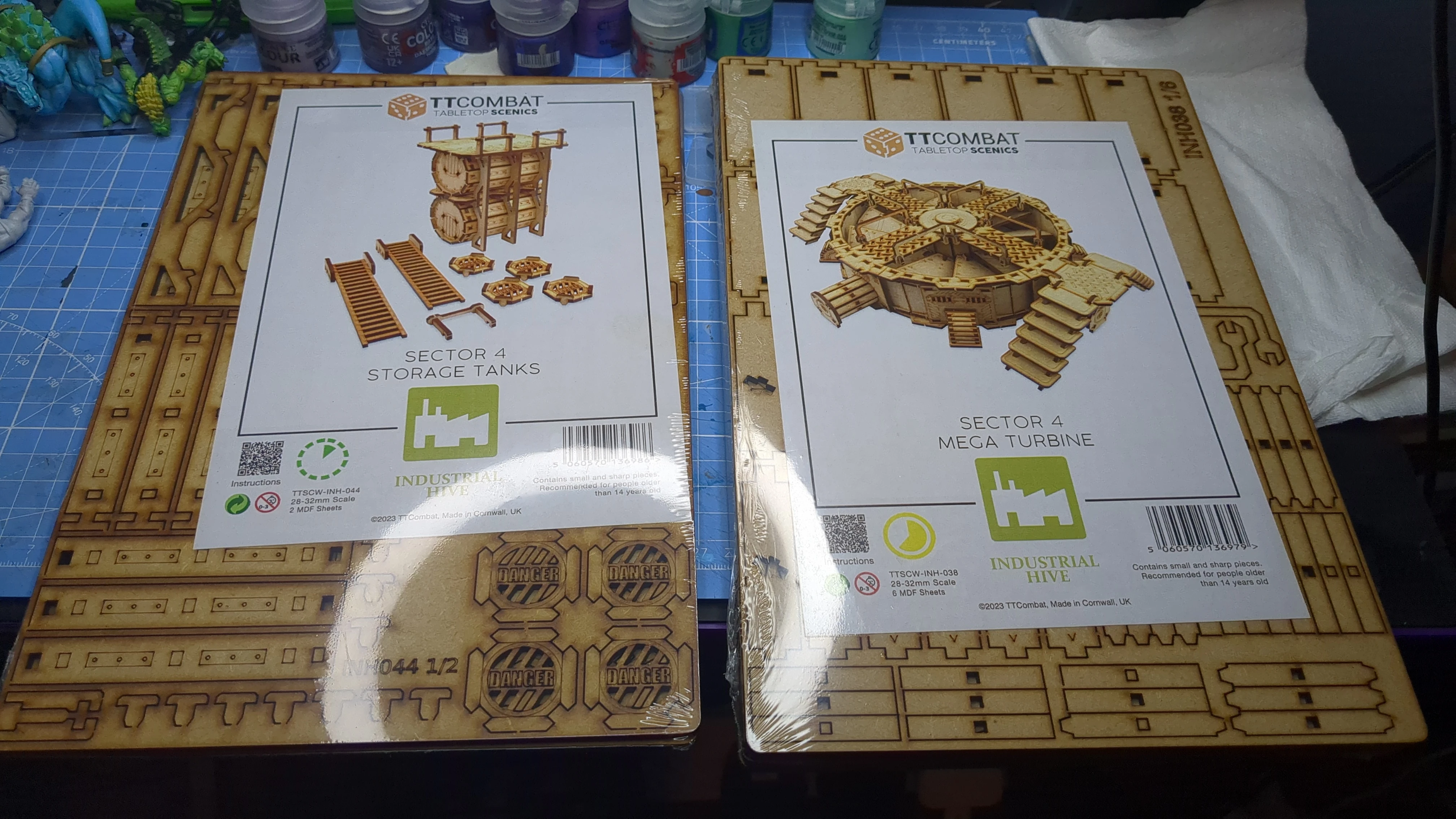 Two boxes of TT Combat tabletop scenery are displayed on a work surface. The left box is labeled "Sector 4 Storage Tanks," featuring images of storage tanks, ladders, and crates. The right box is labeled "Sector 4 Mega Turbine," showcasing a detailed turbine design. In the background, various paint bottles are visible, indicating a workspace for model building and painting.