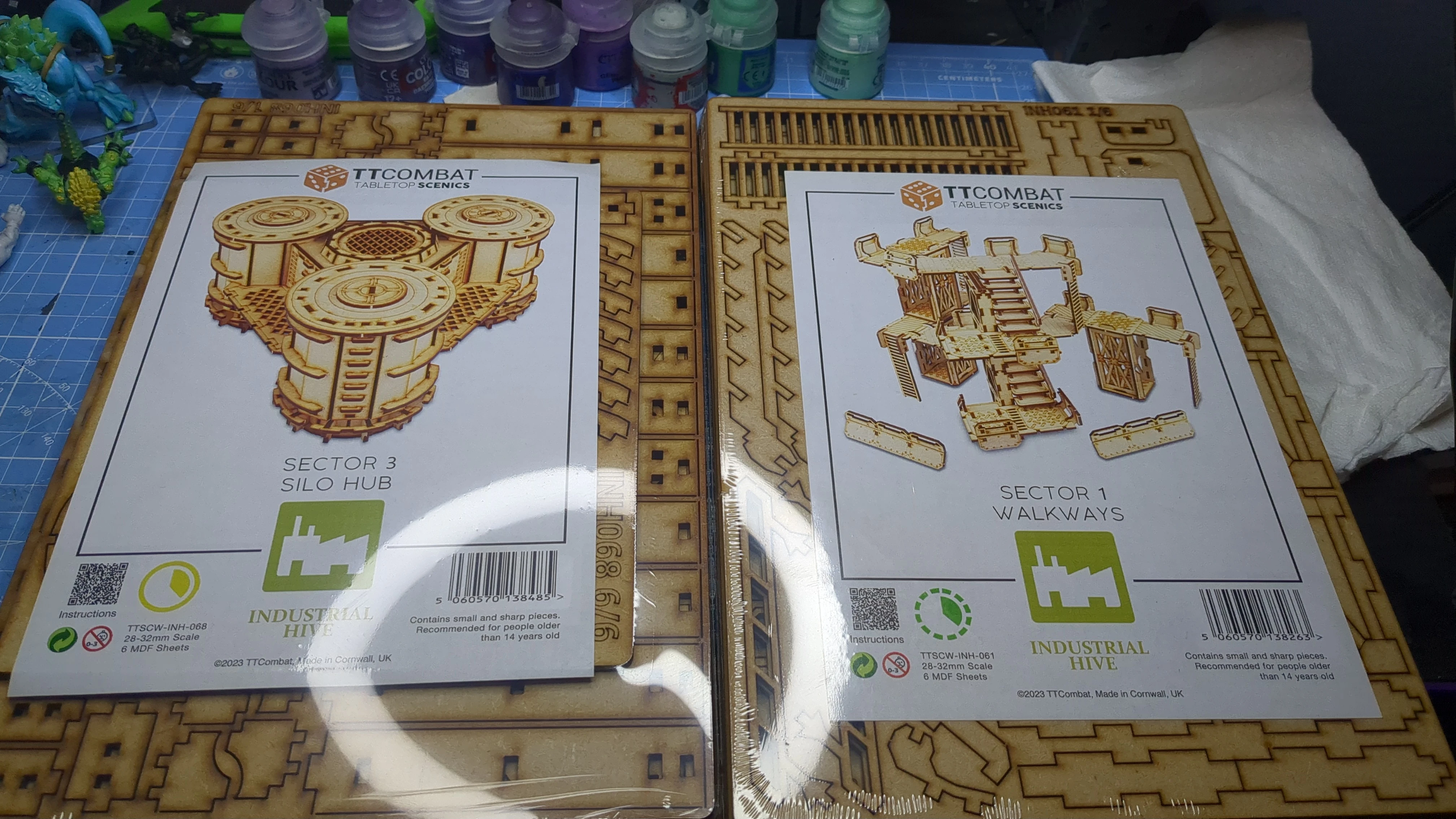 Two packages of TT Combat tabletop scenery are displayed on a work surface. On the left, the "Sector 3 Hub" features detailed illustrations of storage tanks, while the right package showcases "Sector 1 Walkways" with images of a multi-level structure and stairs. In the background, various paint bottles in shades of green and purple are visible, indicating preparation for painting the models.