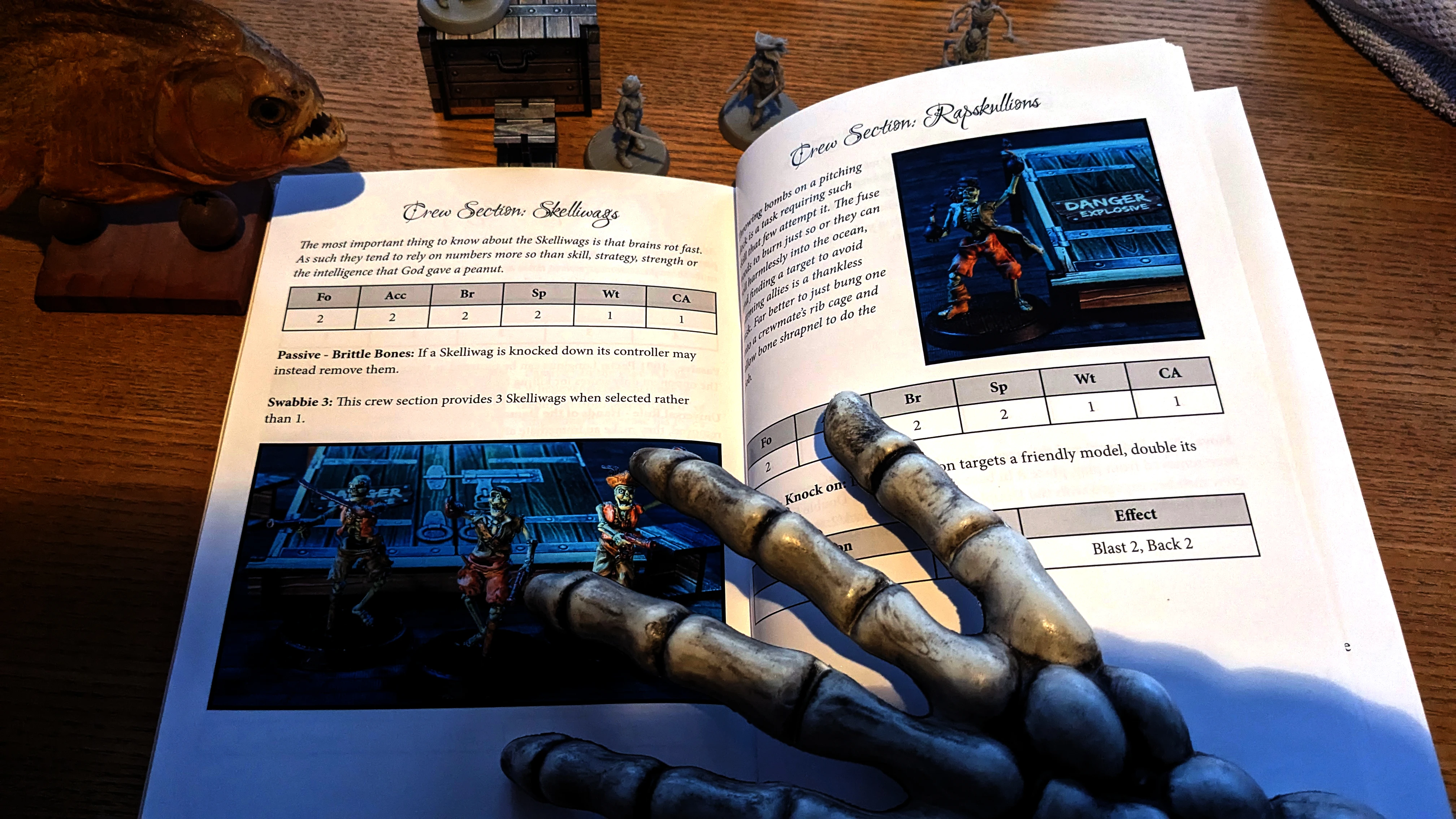 A tabletop scene featuring a partially open rulebook for the game "Boarders and Black Flags," displaying sections on Skelliwags and Rapskullions, with accompanying illustrations of pirate miniatures. In the foreground, a skeletal hand is positioned, adding a thematic touch to the pirate aesthetic. Behind the book, several pirate miniatures are arranged, showcasing their detailed sculpts, including a mix of human and undead figures.