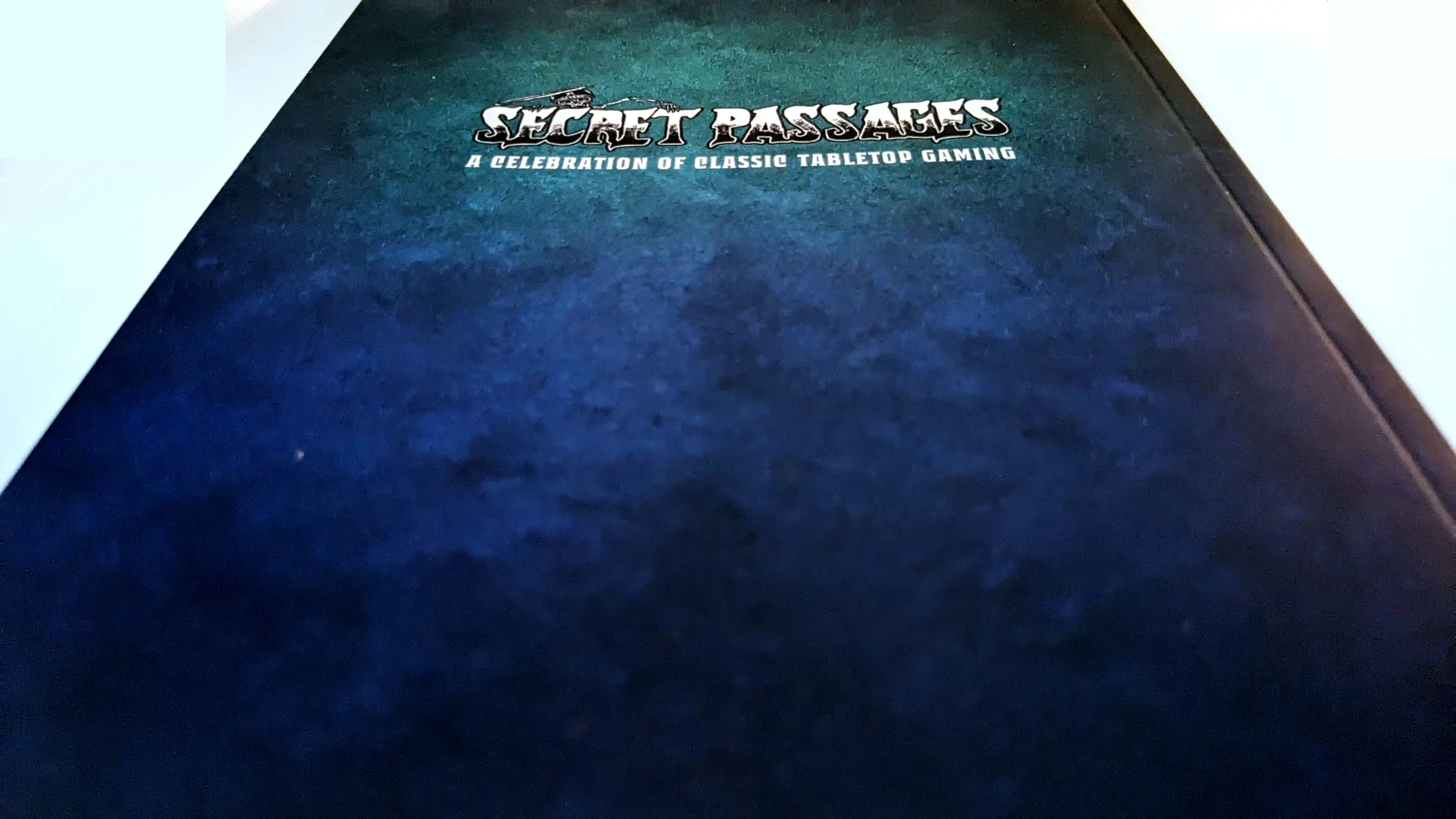 A dark blue cover features the title "SECRET PASSAGES" prominently displayed in a bold, stylized font, with the subtitle "A Celebration of Classic Tabletop Gaming" beneath it. The background has a textured appearance, enhancing the overall aesthetic of the design. The cover suggests a focus on nostalgia and the rich history of tabletop gaming.