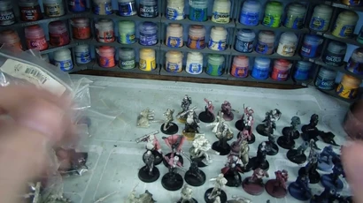 A variety of Heresy Miniatures models are arranged on a work surface, showcasing a mix of painted and unpainted figures in various poses. In the background, shelves filled with paint pots in a range of colors, including blues, greens, and reds, provide a vibrant backdrop. The models include soldiers and fantastical characters, some with detailed bases, while the surface beneath them is marked with paint splatters and tools of the hobby.