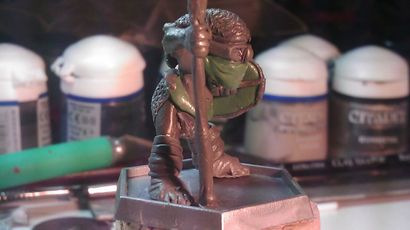 A small, detailed miniature figure stands on a square base, holding a staff and wearing a green mask. The figure is dressed in textured armor, showcasing intricate details like scales and straps. In the background, various paint pots and tools are visible, indicating a workspace for painting or modeling miniatures.