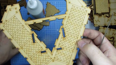 A close-up view of a partially assembled model from the Slum Shanties set by TT Combat, showcasing intricate details like diamond plate textures on the wooden pieces. In the foreground, a hand holds a section of the model, while various cut-out pieces are visible on a blue cutting mat in the background. A bottle of glue is also present, indicating the assembly process.