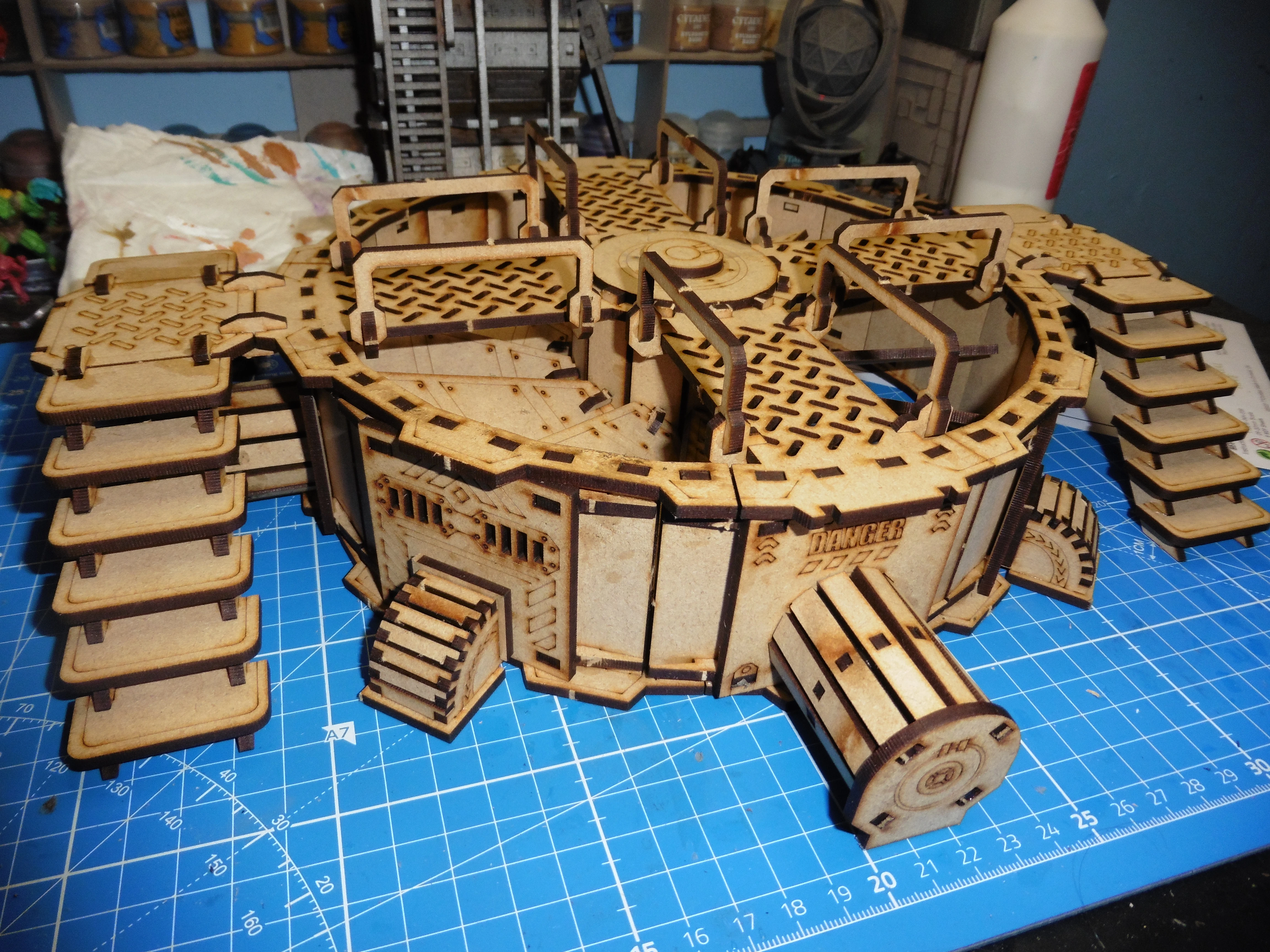 A detailed model of the TTCombat Mega Turbine, featuring a circular base with multiple levels and intricate cut-out patterns. The structure includes staircases leading up to the main platform, which is adorned with various mechanical details and a central turbine section. The model is set against a blue cutting mat, with tools and paint supplies visible in the background, indicating an active workspace.