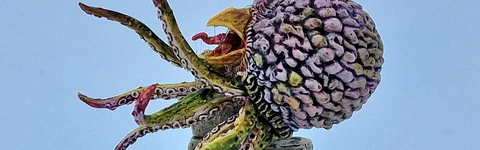 A detailed miniature of a creature resembling a large, bulbous head with a textured surface, featuring vibrant colors like purple and green. It has multiple long, tentacle-like appendages extending from its base, which is perched atop a pile of irregularly shaped stone blocks. The base is circular and textured, resembling a rocky terrain, enhancing the overall fantasy aesthetic of the piece.