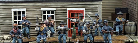 A group of ten detailed 28mm scale miniatures representing dismounted US Cavalry and Buffalo Soldiers stands in front of a wooden building with a red door. The miniatures are posed with various weapons, including rifles and a bugle, showcasing a mix of uniforms and accessories, such as scarves and hats. A small beagle and a cactus are also present among the figures, all mounted on 25mm round bases with a sandy terrain effect.
