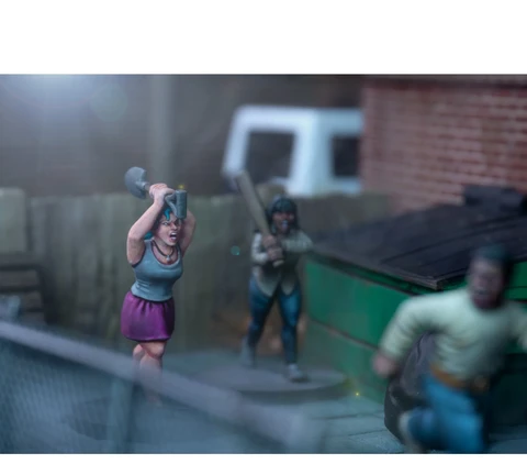 A dynamic scene features three miniature figures in a tense urban setting. In the foreground, a woman with blue hair raises a shovel, while a second figure behind her holds a baseball bat, both appearing ready to confront a threat. A third figure, dressed in a light shirt and blue pants, is seen running away, adding to the sense of urgency in the scene, with a green dumpster and a wooden fence in the background.