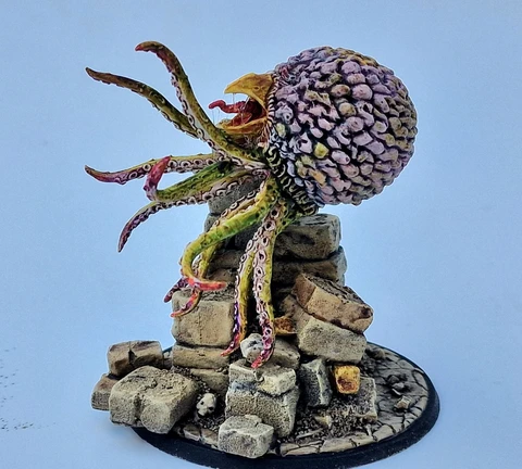 A detailed miniature of a creature resembling a large, bulbous head with a textured surface, featuring vibrant colors like purple and green. It has multiple long, tentacle-like appendages extending from its base, which is perched atop a pile of irregularly shaped stone blocks. The base is circular and textured, resembling a rocky terrain, enhancing the overall fantasy aesthetic of the piece.