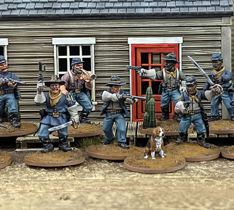 A group of ten detailed 28mm scale miniatures representing dismounted US Cavalry and Buffalo Soldiers stands in front of a wooden building with a red door. The miniatures are posed with various weapons, including rifles and a bugle, showcasing a mix of uniforms and accessories, such as scarves and hats. A small beagle and a cactus are also present among the figures, all mounted on 25mm round bases with a sandy terrain effect.