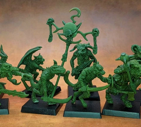 A collection of old school miniatures from Satyr Art Studio, featuring various fantastical creatures in a vibrant green color. The figures include beastmen with distinct features such as horns and claws, some wielding weapons, and a central figure holding a staff topped with a skull and a crescent. They are displayed on black bases against a warm, textured background.