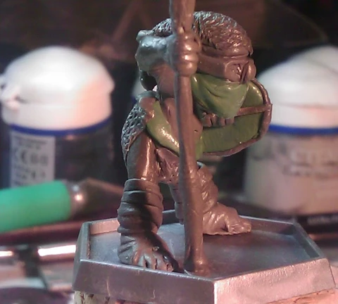 A small, detailed miniature figure stands on a square base, holding a staff and wearing a green mask. The figure is dressed in textured armor, showcasing intricate details like scales and straps. In the background, various paint pots and tools are visible, indicating a workspace for painting or modeling miniatures.