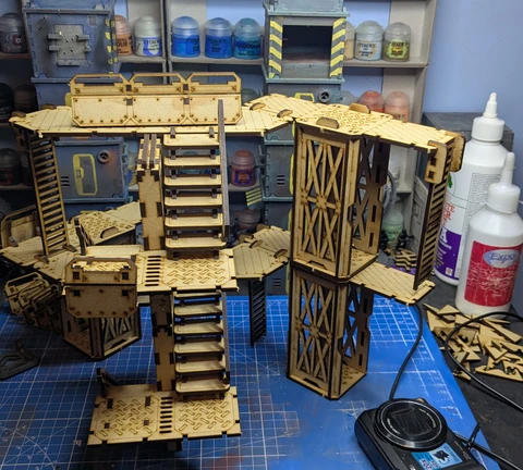A detailed assembly of modular terrain pieces made from laser-cut wood, featuring multiple levels, staircases, and platforms. The structure includes various textures and patterns, with a mix of open and enclosed areas, showcasing a futuristic industrial design. In the background, paint bottles and tools are visible, indicating a workspace for painting and customizing the terrain.