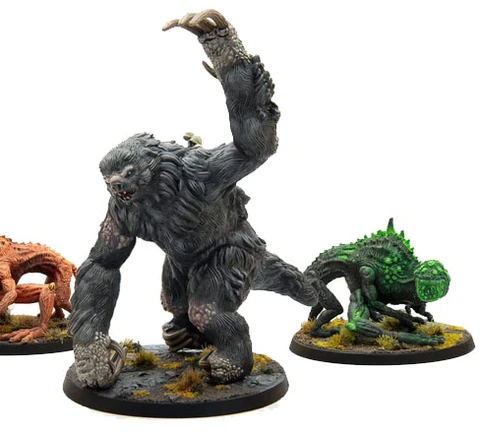 The image features three miniatures from Modiphius’ Fallout: Wasteland Warfare line. On the left is a snallygaster, a hexapod creature with a reddish-brown body, multiple eyes on its back, and a long, protruding tongue. In the center stands a large, muscular megasloth, covered in gray fur, with a menacing pose and a raised arm. To the right is another creature, possibly a variant of the snallygaster, in green, with a textured body and large eyes, all set on circular bases adorned with tufts of grass.