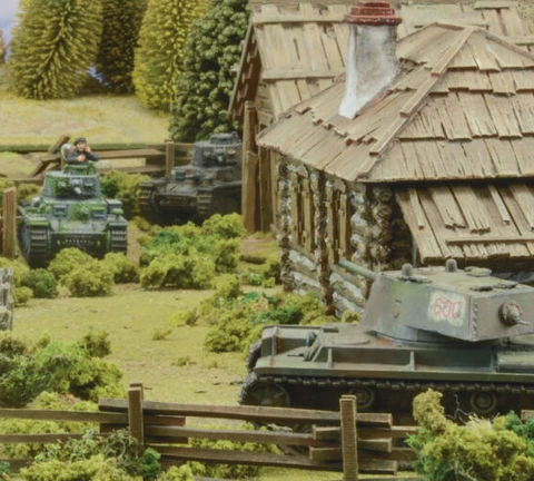 A detailed tabletop scene features several miniature tanks amidst a rural setting, including a wooden cabin and lush greenery. In the foreground, a tank with the number "600" is positioned near a wooden fence, while another tank is seen further back with a crew member visible. The background includes trees and additional structures, creating a vibrant battlefield atmosphere.