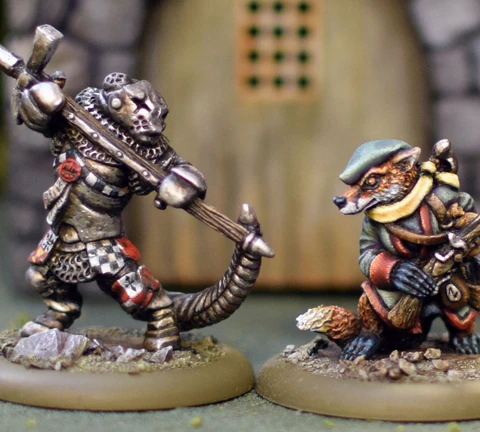 A fierce anthropomorphic badger in armor wields a large weapon, standing on the left, while a cunning-looking fox in a green hat and coat stands on the right, holding a small object. Both miniatures are detailed and painted, set against a blurred background of a stone structure with a wooden door. The scene captures the whimsical yet combative spirit of the Burrows and Badgers game.