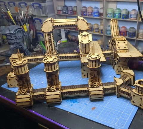 A detailed assembly of the TT Combat Sector 4 Plasma Relay is displayed, featuring intricate wooden structures with multiple towers and connecting pathways. The design includes cylindrical towers with circular tops and various platforms, all set against a blue cutting mat. In the background, jars of paint and tools are visible, indicating a workspace dedicated to tabletop gaming and model building.
