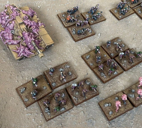 A collection of Zayken miniatures from Void Scar Miniatures is displayed on a textured surface. In the foreground, several bases feature hordes of zombified figures in various colors, including green, purple, and pink, representing the Neophyte Mobs. To the left, a tank is adorned with grotesque, bloated creatures, suggesting a fusion of machine and undead, while additional bases of zombies are scattered throughout the scene, emphasizing the overwhelming numbers of the Zayken forces.