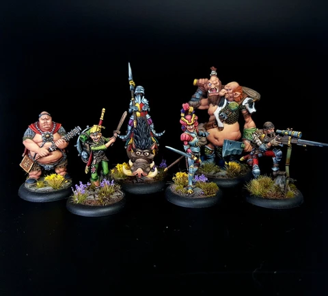 A group of eight intricately painted Moonstone miniatures stands against a black background. The characters include a muscular figure with a weapon, a green-skinned goblin in colorful attire, a knight in armor, and various other fantastical beings, each displaying unique features and vibrant colors. They are positioned on circular bases adorned with tufts of grass and small flowers, showcasing the detailed artistry of the miniatures.