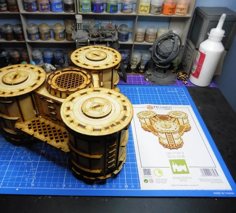 A detailed model of the Sector 3 Silo Hub from TT Combat is prominently displayed on a blue cutting mat. The structure features multiple cylindrical silos with intricate wooden textures and circular tops, showcasing a combination of open latticework and solid panels. In the background, various paint pots and a few other miniature models are visible, along with the instruction sheet for the Silo Hub, which includes an illustration of the assembled model.