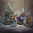 Four intricately designed miniatures from the underwater tabletop skirmish game DeepWars are displayed against a neutral background. On the left, a large, armored figure with a green and blue color scheme wields a large ice sword. Next to it, a humanoid character in vibrant red and green attire gestures dramatically, showcasing blue skin and a confident expression. The third figure, a mermaid-like character with flowing purple hair and a shimmering tail, holds a staff adorned with a glowing orb. On the right, a warrior with a blue head and patterned armor brandishes a large axe, completing the diverse ensemble of characters. Each miniature is mounted on a textured base that reflects an underwater theme.