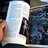 A hand holds open a page from the publication "Secret Passages," revealing text discussing character creation in the context of the game "Inquisitor." On the left side, there is an illustration of a character labeled "The Harlequin," depicted in a stylized, jester-like costume. The right side features an advertisement for Fenris Games, showcasing a variety of miniatures and scenery, with vibrant colors and intricate details. The background suggests a cozy indoor setting, with a wooden table surface visible.