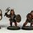 Three fantasy miniatures are displayed, each depicting a character in armor with distinct features. The first figure on the left holds a bow, while the middle figure wields an axe and a shield, showcasing a fierce expression. The third figure on the right is armed with a spear, all standing on textured bases with a hint of grass. Each miniature is painted with vibrant colors, emphasizing their detailed armor and facial expressions.