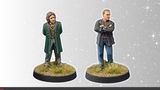 A pair of miniature figures titled "Temporal Travellers" stands on a textured base with tufts of grass. On the left, a figure in a green coat and vest has curly hair and a confident stance, while on the right, a figure in a black jacket with crossed arms and short hair presents a more serious demeanor. The background features a subtle starry effect, enhancing the theme of time travel.