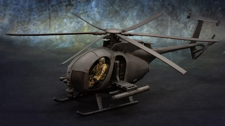 A detailed model of a Little Bird helicopter, specifically the AH-6 variant, is prominently displayed. The helicopter features a sleek, dark exterior and is equipped with a mounted weapon system on the side. Inside the cockpit, a miniature figure dressed in camouflage is seated, showcasing the model's intricate design and attention to detail. The background is a textured gradient, enhancing the focus on the helicopter.