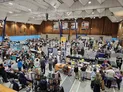 Indoor event hall with high ceiling and wooden walls. Crowded with attendees walking and browsing. Multiple vendor tables displaying board games, books, and gaming accessories. Banners and posters on walls, including one with "Paradice" and another with "Gamer’s Zone."