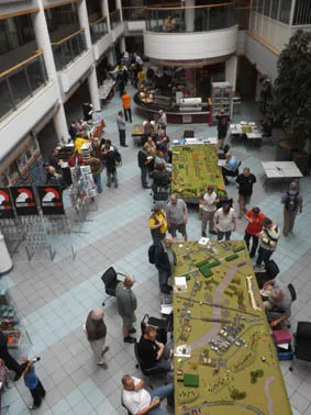 A bustling convention scene at Sheffield Hallam University, featuring a large gaming table covered with a green terrain layout, complete with miniature buildings and vehicles. Attendees, dressed in casual clothing, are engaged in various activities around the table, while others browse nearby displays. The open space is bright and airy, with a café area visible in the background.