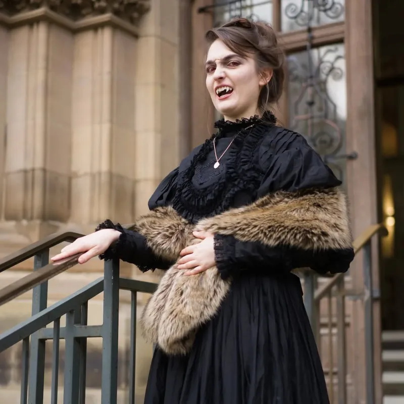 A person dressed in a gothic-style black dress stands on a staircase, exuding a vampire aesthetic with fangs visible and a playful expression. They are adorned with a fur stole draped over their shoulders and a necklace, enhancing the dramatic look. The backdrop features the ornate architecture of a historic building, contributing to the overall atmospheric setting.