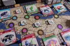 A board game with a map featuring dotted paths and terrain. Four character cards labeled "Björn," "Tarmaby," "Are," and "Linné" with illustrations and text. Various colored tokens, cards with icons, and numbered dice on the board.
