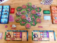 Board game setup with hexagonal green terrain tiles featuring pink and purple structures. Miniature figures and small tokens scattered across. Two player boards with character portraits, resource dials, and action icons. Dice and small bags of colored tokens on the table.