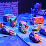 A vibrant tabletop scene features a variety of uniquely designed miniature vehicles, each with whimsical and chaotic elements, such as spikes, exaggerated features, and colorful paint jobs. The vehicles are arranged on a textured surface that resembles a racing track, with a backdrop of stylized scenery, including a stone wall and scattered debris. The lighting adds a dynamic flair, enhancing the fantastical atmosphere of the racing game "Fool's Fuel."