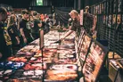 A bustling convention scene filled with attendees exploring various stalls and displays. In the foreground, a long table is covered with vibrant artwork and prints, showcasing horror-themed images, while a person examines the items closely. The background features a crowd of people, some in themed attire, engaging with the immersive atmosphere of the event.