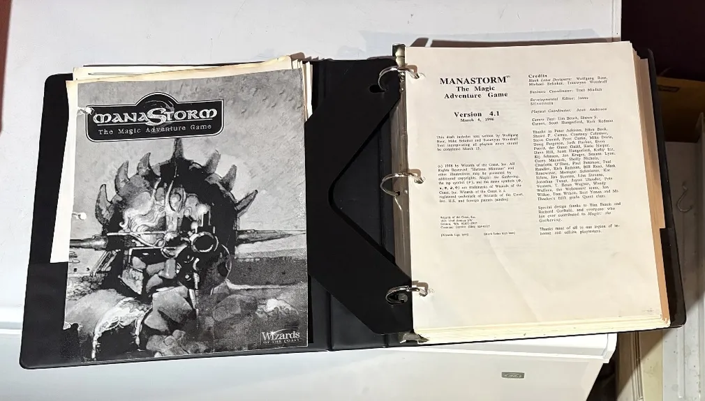 A three-ring binder containing the playtest manuscript for "ManaStorm: The Magic Adventure Game," featuring a black and white cover illustration of a character with a spiked helmet and a weapon. The title "MANASTORM The Magic Adventure Game" is prominently displayed at the top, along with the version number and date on the first page inside the binder. Additional pages include loose character sheets and player handouts, indicating the game's mechanics and spells.