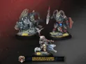 A trio of miniatures from the Khador Old Umbrey Battlegroup Box Set is displayed against a dark, textured background. On the left, a fierce, wolf-like warbeast with spiky fur stands on a rocky base, while in the center, a character wielding a rifle is dressed in fur-lined clothing, poised for action. To the right, a heavily muscled figure with a ram-like head brandishes a spear and shield, adorned with tribal elements. The bottom of the image features a banner with the text "KHADOR OLD UMBREY BATTLEGROUP BOX SET."