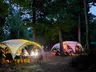 A cozy outdoor gaming setup features several large, white tents illuminated by warm lights, surrounded by trees in a wooded area during dusk. Groups of people are seated at tables under the tents, engaged in games and conversation, creating a relaxed and social atmosphere. The ground is covered in leaves, and the soft glow from the lights adds to the inviting ambiance of the gathering.
