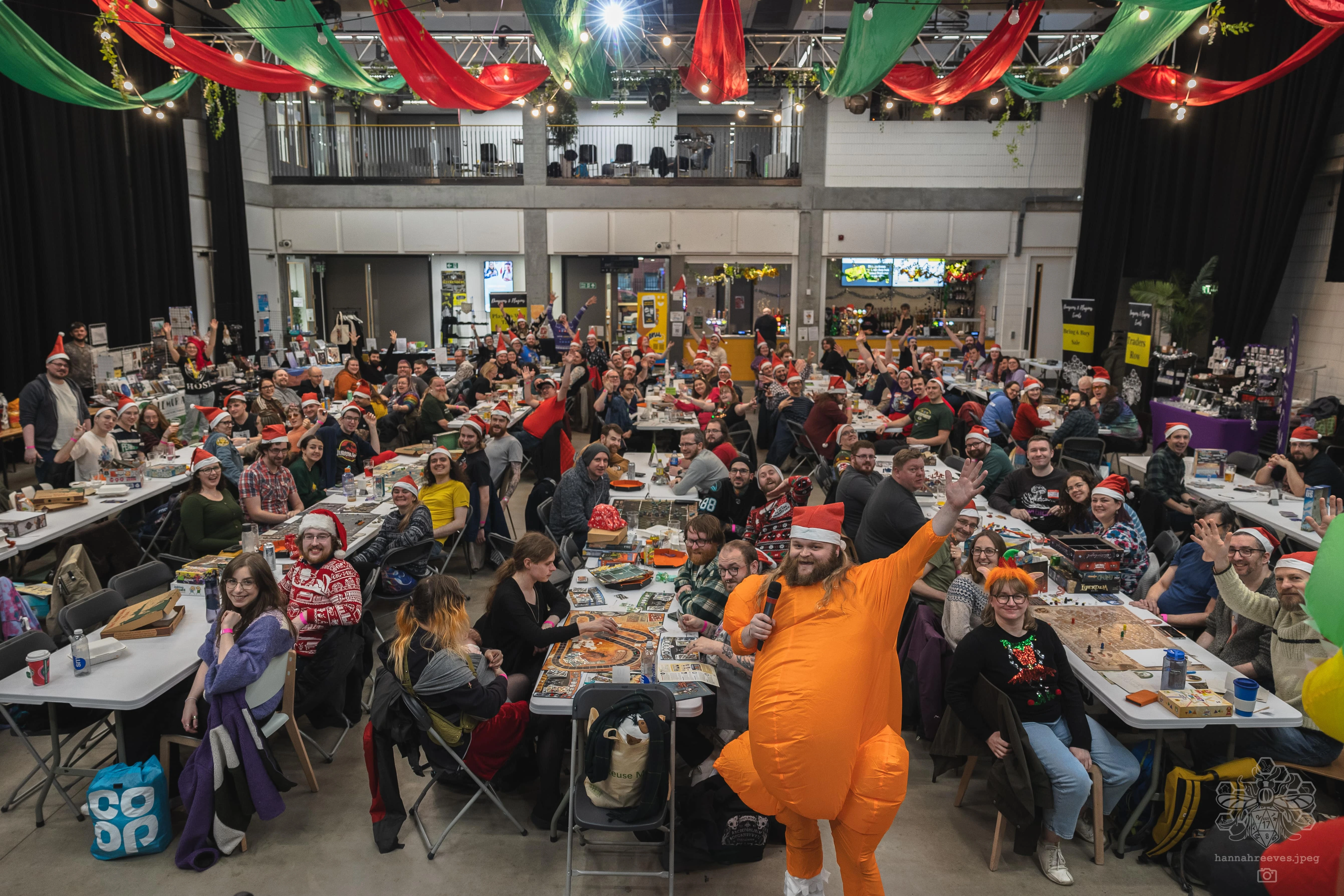 A lively scene captures a large gathering of people in a festive atmosphere, with many attendees wearing Christmas hats and themed attire. In the foreground, a man in an orange inflatable costume holds a microphone, engaging the crowd, while tables filled with board games and drinks stretch into the background. Colorful decorations, including red and green drapes, enhance the celebratory mood of the event.