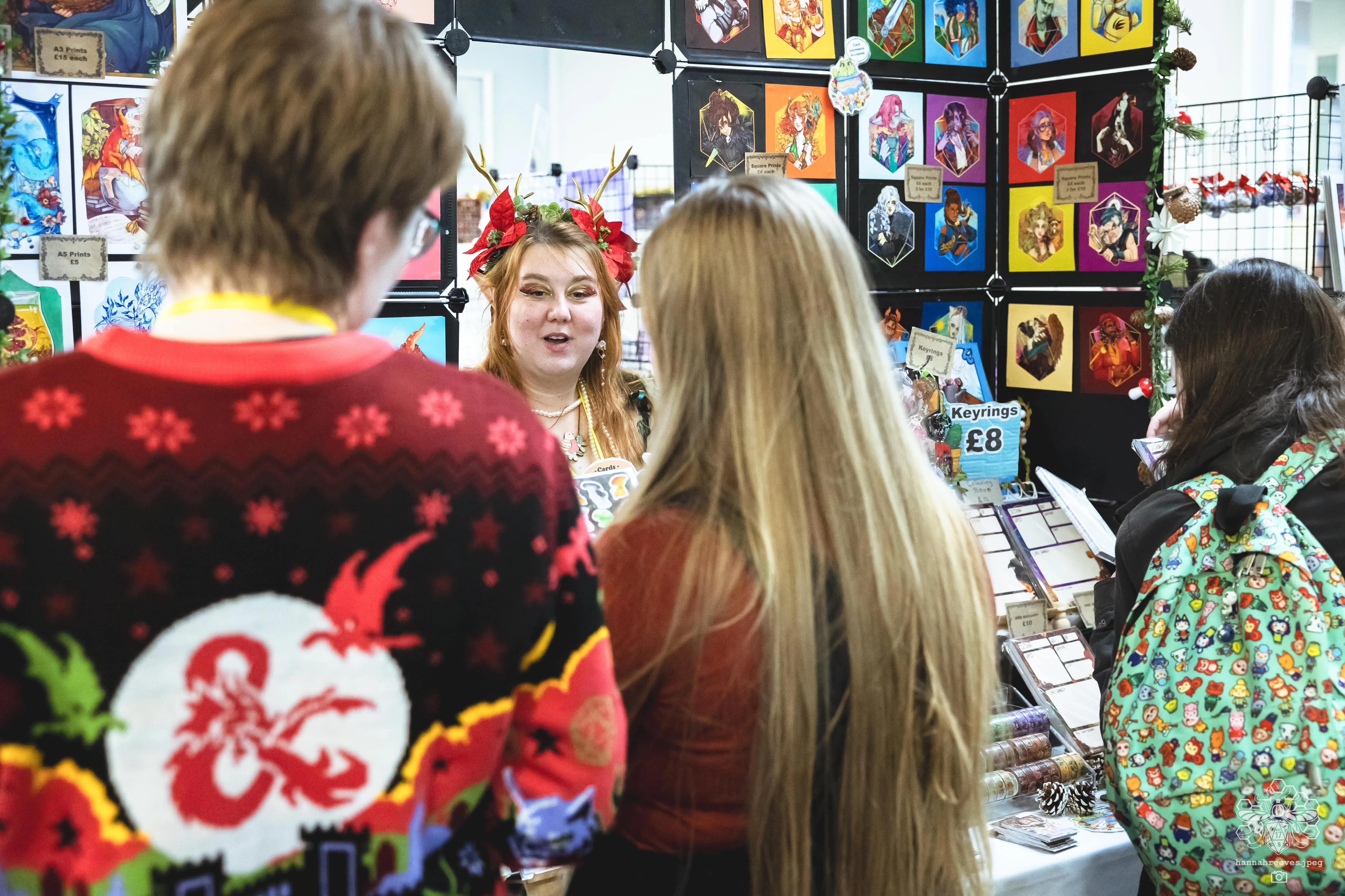 A festive scene at a board game festival features a woman with antlers and floral decorations in her hair, engaging with two attendees. One attendee wears a red sweater adorned with a Dungeons & Dragons logo, while the other has long, straight hair. Behind them, a display showcases various colorful artwork and merchandise, including keyrings for sale.