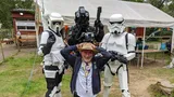 A man in a wide-brimmed hat and a casual outfit kneels in the foreground, smiling and playfully raising his hands above his head. Behind him, two Stormtroopers and a TIE Fighter pilot in full costume stand with their weapons, creating a fun and engaging atmosphere. The setting features a grassy area with a tent in the background, suggesting a lively outdoor event.