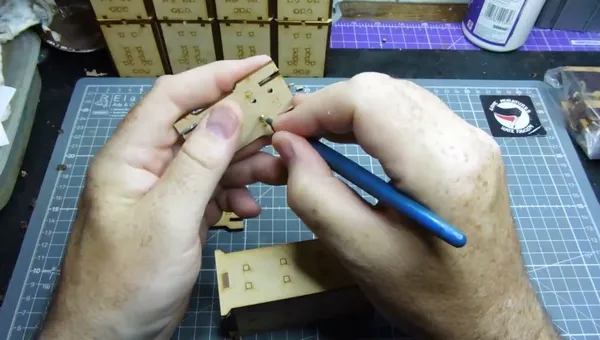Two hands holding a partially assembled laser-cut MDF piece for a miniature building kit. The left hand supports the main structure, which is a yellowish-beige MDF panel with square cutouts and small circular holes. The right hand uses a blue-handled metal tool, likely tweezers or a small pick, to position or secure a small metal disc, possibly a magnet, into one of the circular holes. In the background, a clear plastic cutting mat with a printed grid is visible, along with several other laser-cut MDF sheets still in their frames, some with engraved or printed text and symbols. One of the MDF sheets in the background has a purple grid pattern. No readable text is visible on the main components being handled.