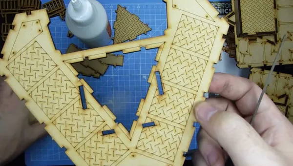 A close-up view of a partially assembled model from the Slum Shanties set by TT Combat, showcasing intricate details like diamond plate textures on the wooden pieces. In the foreground, a hand holds a section of the model, while various cut-out pieces are visible on a blue cutting mat in the background. A bottle of glue is also present, indicating the assembly process.