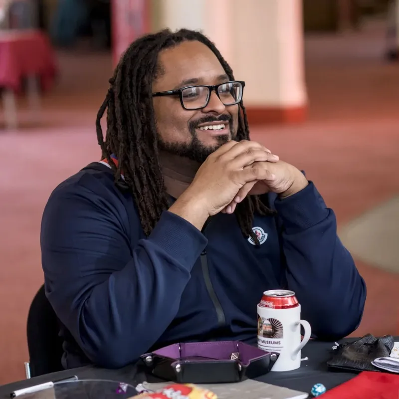 A smiling individual with long, dark hair styled in dreadlocks sits at a table, resting their chin on clasped hands. They are wearing a navy blue jacket and glasses, with a can of soda and a mug featuring a logo on the table in front of them. The background shows a softly lit space, likely part of a larger venue, with hints of red tablecloths and other gaming materials scattered around.